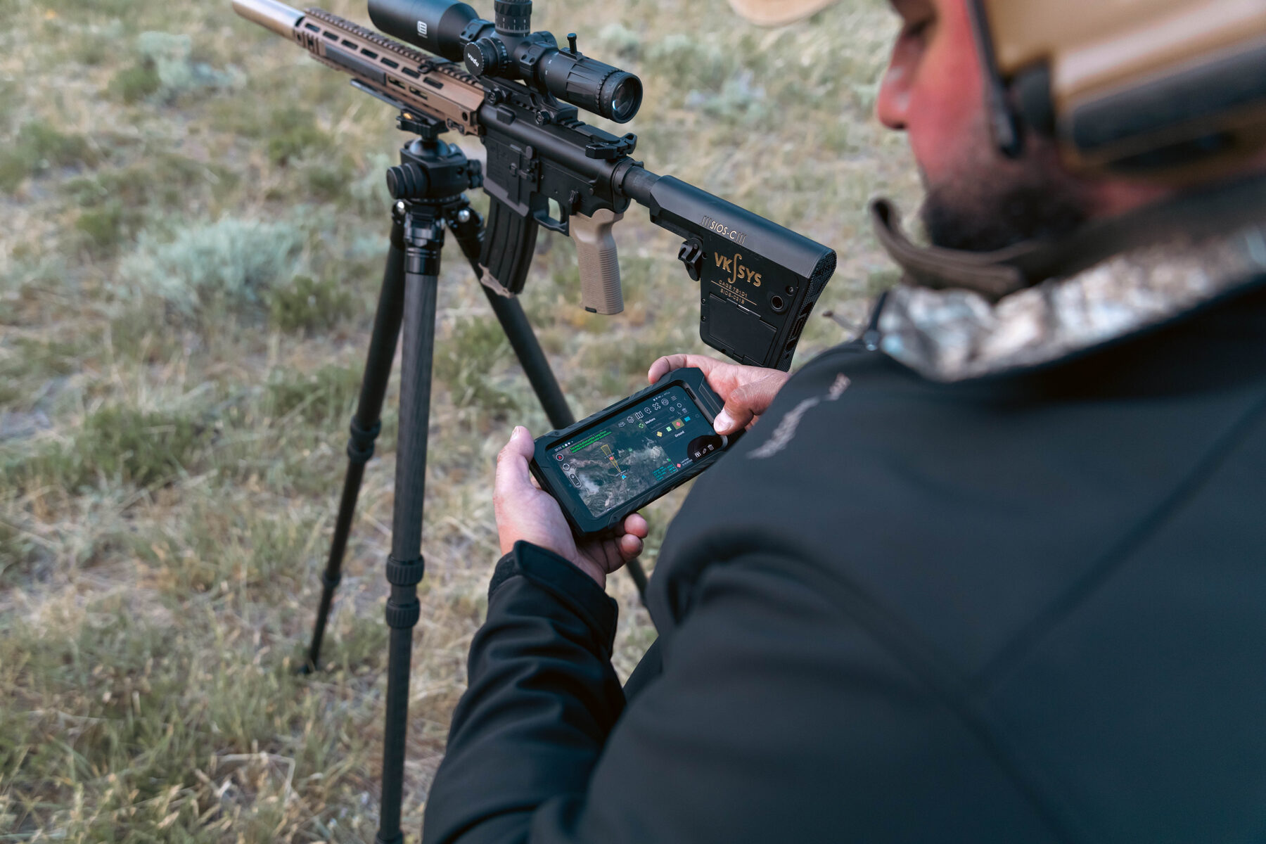 Shooter behind a tripod-mounted rifle holding a phone running ATAK