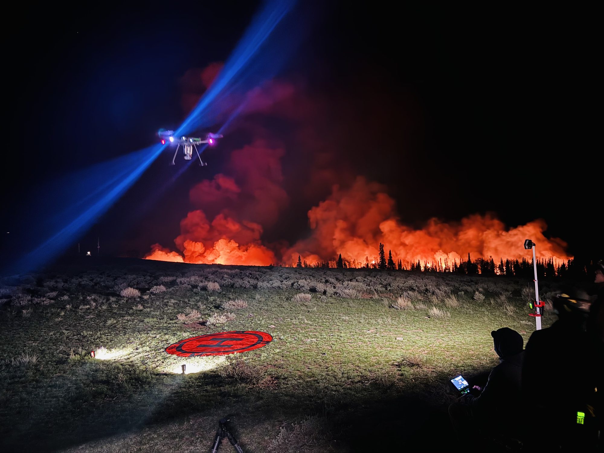 A crew working a night burn from a hilltop, an aircraft above a lit landing pad with the fire front along the treeline beyond